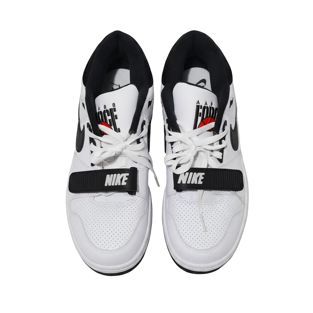 A pair of white and black Nike Low-top sneakers with white laces, displayed on a yellow surface. The shoes feature black Swooshes, black ankle straps with white 'NIKE' text, and a graphic on the tongue.