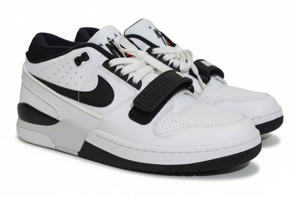 A pair of Nike low-top sneakers in white and black, featuring a black hook-and-loop strap across the laces, photographed on an orange surface against a light gray background.