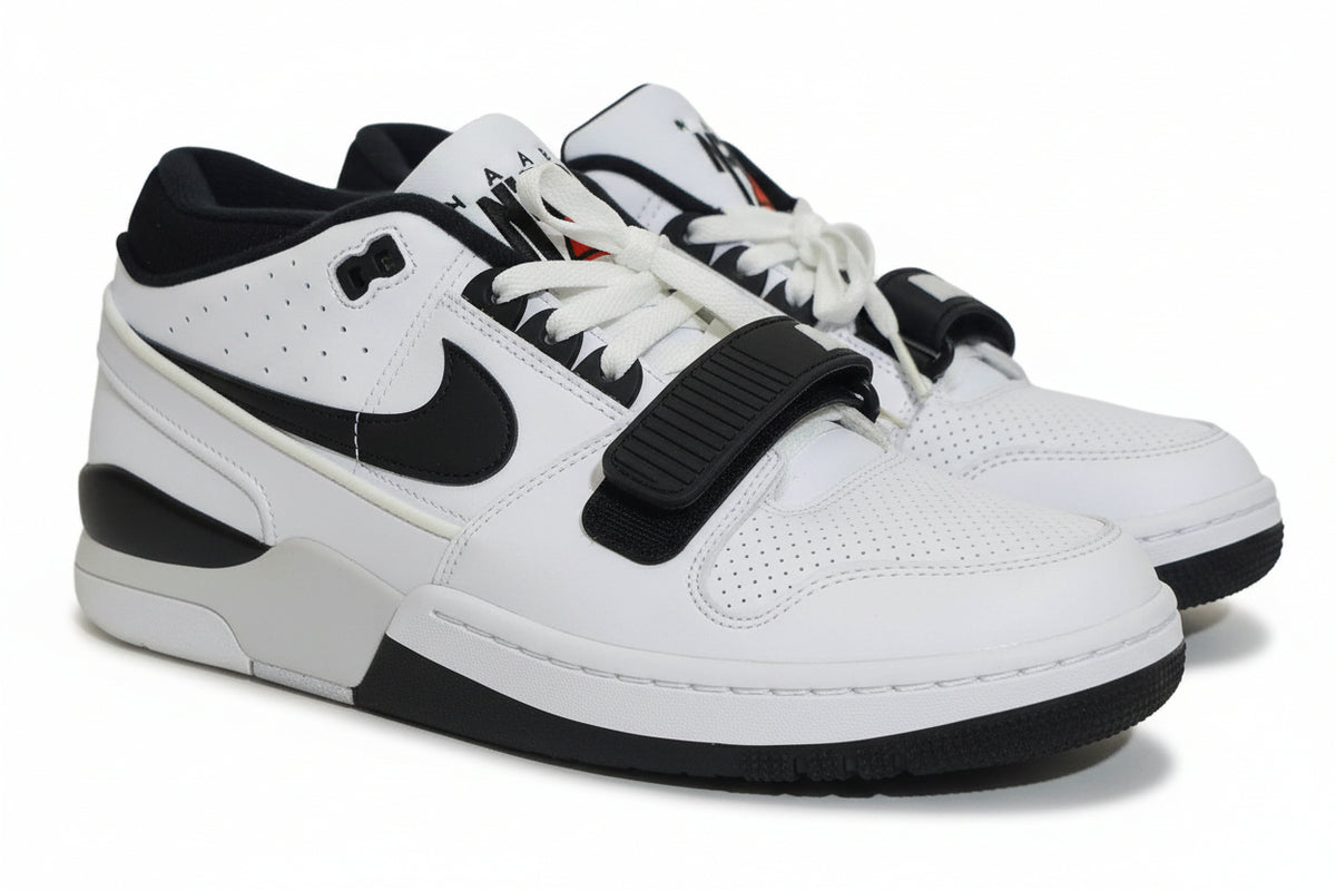 A pair of Nike low-top sneakers in white and black, featuring a black hook-and-loop strap across the laces, photographed on an orange surface against a light gray background.