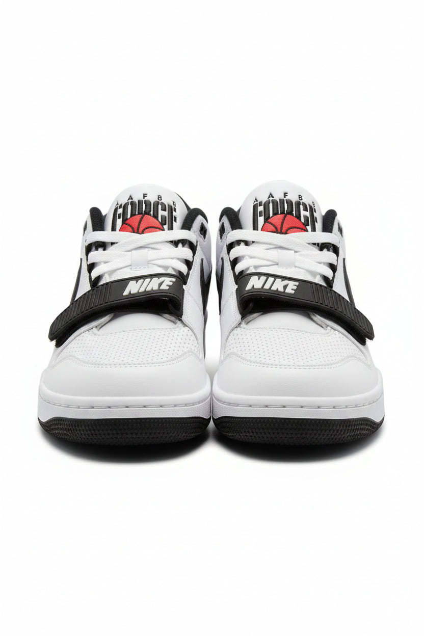 A pair of Nike low-top sneakers in white, black, and red, featuring a black hook-and-loop strap with white 'NIKE' text across the front and a red basketball graphic on the tongue. The shoes are photographed on an orange surface with a beige background.