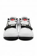 A pair of Nike low-top sneakers in white, black, and red, featuring a black hook-and-loop strap with white 'NIKE' text across the front and a red basketball graphic on the tongue. The shoes are photographed on an orange surface with a beige background.