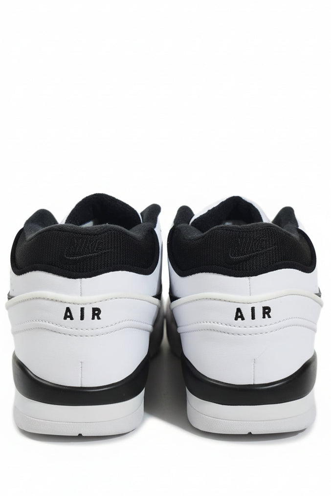 A pair of Nike low-top sneakers shown from the back, featuring a white leather upper with black knit collar, black 'AIR' text, and black midsoles, set against an orange surface with a light beige background.